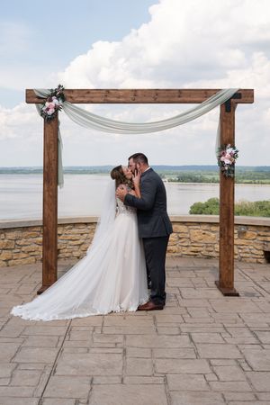 Bride and groom share first kiss under rustic wooden arch with floral accents and river backdrop on stone patio.
