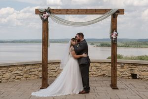 Newlyweds kiss under wooden wedding arch decorated with pink florals and white draping with lake backdrop.