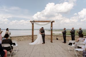 Bride and groom share first kiss under wooden arch at outdoor waterfront wedding ceremony with guests seated.