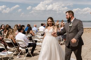 Smiling newlywed couple walks hand in hand down the aisle after outdoor lakeside wedding ceremony.