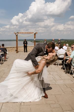Groom dips bride for kiss after outdoor wedding ceremony with scenic lake view backdrop.