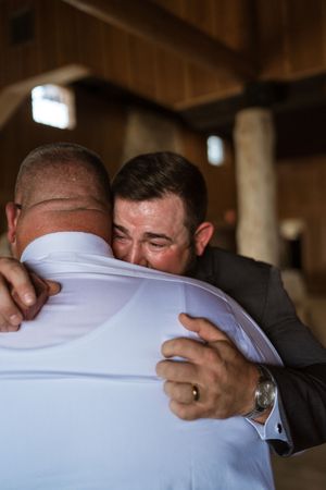 Emotional groom shares heartfelt hug with family member inside rustic wedding venue.