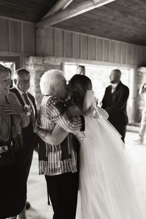 Black and white photo of a bride in a white gown receiving a kiss from an elderly woman at a wedding reception.