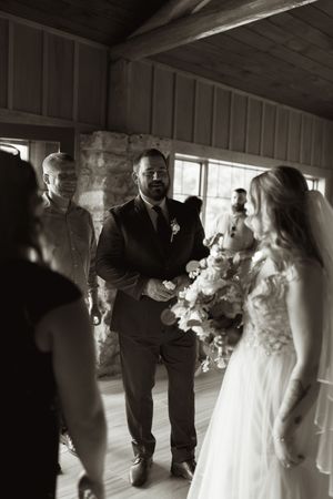Black and white photo of groom in dark suit with boutonniere meeting bride at wedding ceremony indoor venue.