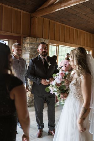 Groom smiling at bride during indoor wedding ceremony in rustic wood-paneled venue with stone fireplace.