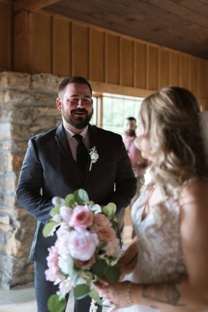 Groom in dark suit smiling at bride holding pink floral bouquet during wedding first look inside rustic wood venue.