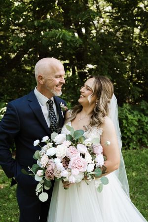 Smiling bride and groom pose together outdoors holding pink and white wedding bouquet.