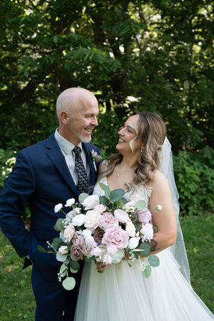 Bride and bald groom in navy suit smiling at each other holding pink and white floral bouquet.