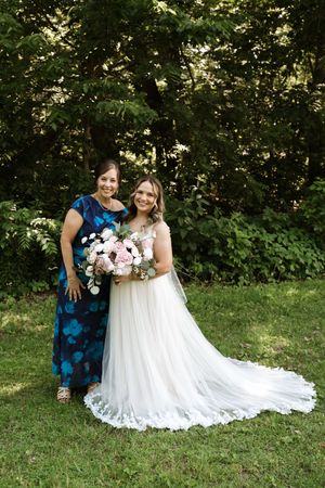Bride in white gown holding pink bouquet poses with woman in blue floral dress outdoors.