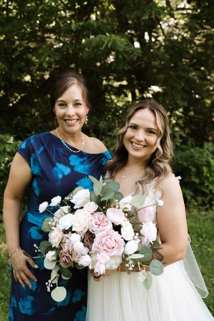 Bride in white gown holds pink bouquet beside woman in blue dress outdoors.