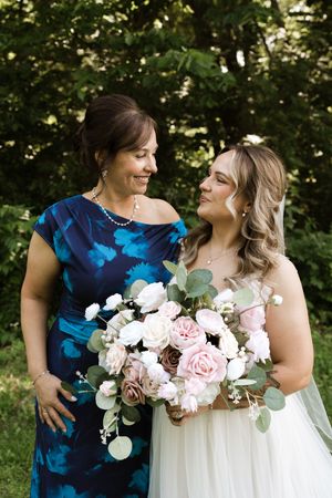 Bride in white gown holding pink and white bouquet smiles with woman in blue floral dress outdoors.