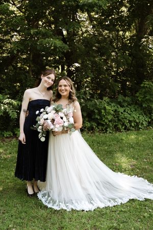 Bride in white gown holds pink bouquet beside bridesmaid in black dress outdoors.