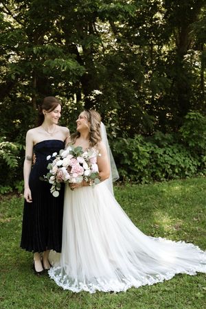 Bride in white lace gown holding pink bouquet smiling with bridesmaid in black dress outdoors.