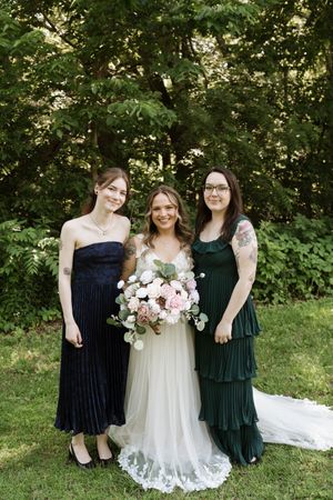 Bride in white gown holding bouquet stands with two bridesmaids in dark dresses outdoors.