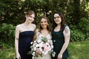 Bride holding pink floral bouquet poses with two bridesmaids in navy and green dresses outdoors.