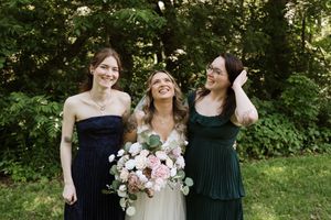 Bride laughing with two bridesmaids in navy and green dresses outdoors in garden setting.