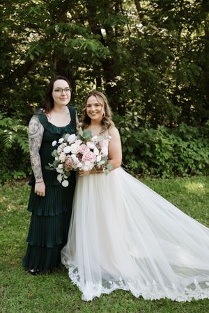 Bride in tulle gown holds floral bouquet with tattooed woman in dark green dress.