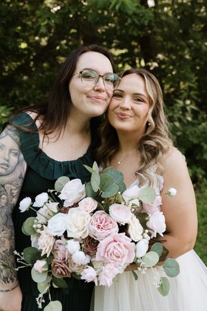 Bride in white gown holds pink floral bouquet with tattooed bridesmaid in green dress, posing outdoors.