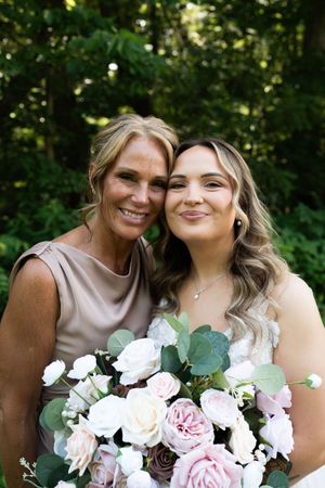 Bride and older woman posing together outdoors with lush green trees, holding pink and white rose bouquet.