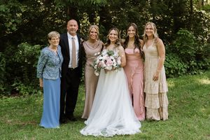 Bride in white gown poses with family and bridesmaids in pink and gold dresses outdoors at a wedding.