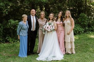 Wedding party poses outdoors, bride in white gown holds pink bouquet, flanked by family in blue, tan, and pink dresses.