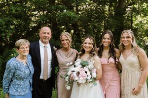 Wedding party group photo with bride holding bouquet, groom, and bridesmaids in pink and gold dresses outdoors.