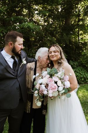 Groom in gray suit and bride in white gown embrace elderly woman between them outdoors.