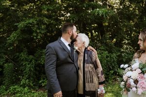 Groom in gray suit kisses elderly grandmother on cheek at outdoor wedding ceremony surrounded by lush green trees.