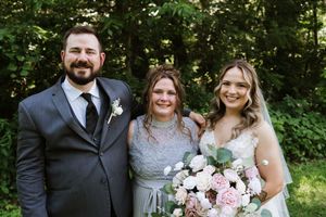 Bride in white gown holding pink bouquet poses with groom in gray suit and bridesmaid in silver dress outdoors.
