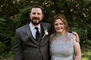 Smiling couple in wedding attire pose together outdoors with lush green trees behind them.