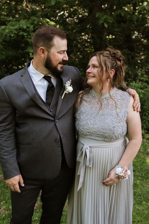 Groom in gray suit with boutonniere smiles at bridesmaid in sage green gown outdoors.