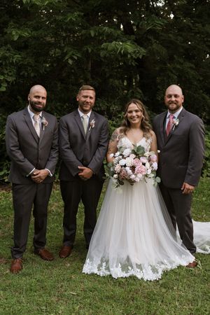 Bride in lace wedding gown holds pink bouquet with three groomsmen in gray suits outdoors.