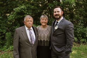 Elderly couple poses with bearded groom in gray suit at outdoor wedding celebration.