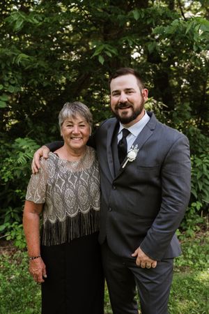 Groom in gray suit with boutonniere poses with smiling older woman in lace top at outdoor wedding.