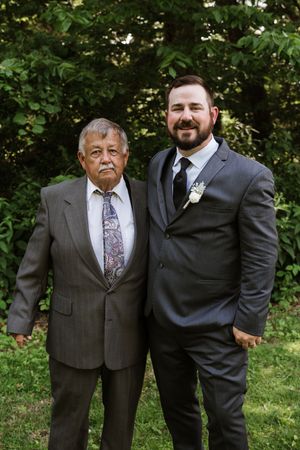 Groom with boutonniere stands beside older man in gray suits outdoors at wedding.