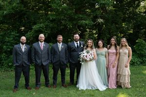Wedding party poses outdoors with groom, groomsmen in gray suits, bride in white gown, and bridesmaids in pastel dresses.