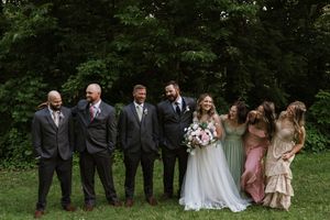 Wedding party poses outdoors with bride in white gown, groom, groomsmen in gray suits, and bridesmaids in sage green dresses.