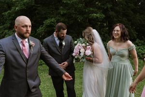 Bride holding pink bouquet laughs with bridesmaid while groomsmen stand nearby at outdoor wedding ceremony.