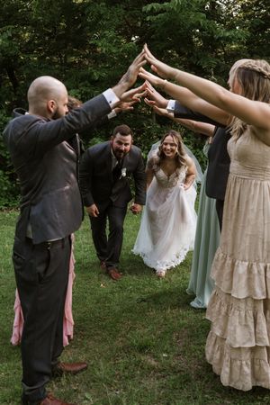 Bride and groom walk through an arch of raised hands formed by smiling wedding party members outdoors.