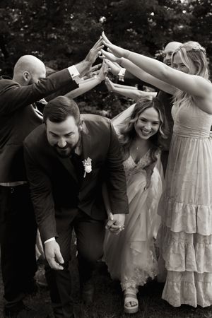Black and white photo of bride and groom walking under an arch of raised hands formed by smiling wedding party members outdoors.