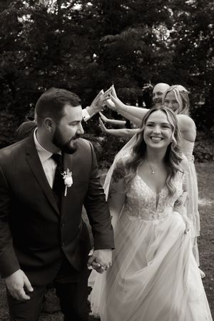 Black and white photo of smiling bride in lace gown holding hands with bearded groom at outdoor wedding.
