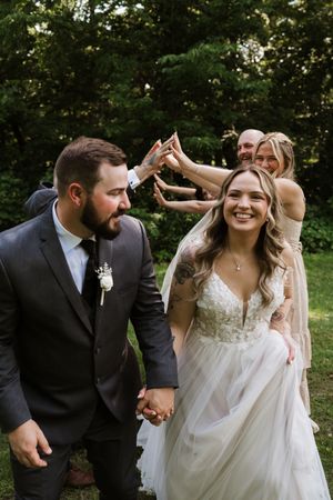 Smiling bride and bearded groom hold hands while wedding party celebrates behind them.