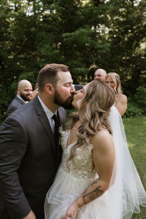 Bride and groom share a kiss surrounded by wedding party in outdoor garden setting.