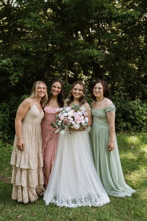 Bride with pink and white bouquet poses with three bridesmaids in mismatched pastel gowns outdoors.