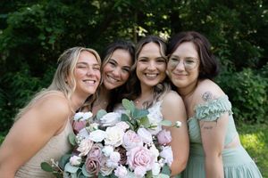 Bride poses with three bridesmaids in sage green dresses, holding pink and white floral bouquet outdoors.