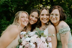 Four bridesmaids in sage green dresses smiling together outdoors holding pink and white floral bouquet.