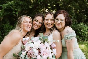 Four bridesmaids in sage green dresses smiling together outdoors holding a pink and white floral bouquet.