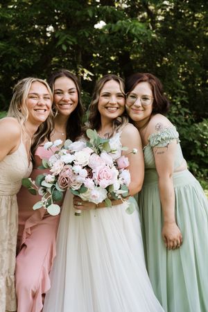 Bride with pink bouquet smiles with four bridesmaids in sage, pink, and gold dresses outdoors.