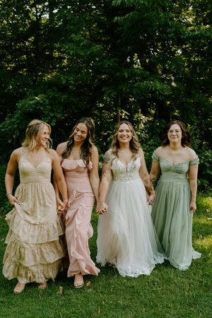 Bride in white tulle gown walks hand-in-hand with three bridesmaids in mismatched pastel dresses outdoors.