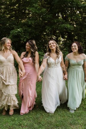 Bride and three bridesmaids in colorful gowns hold hands walking together outdoors.